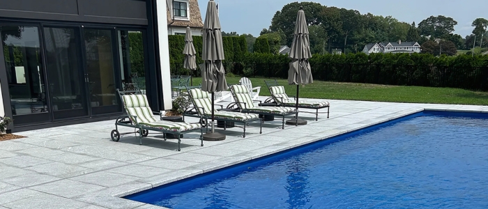 A serene pool with lounge chairs and umbrellas, set on a stone deck in front of a beautiful house.
