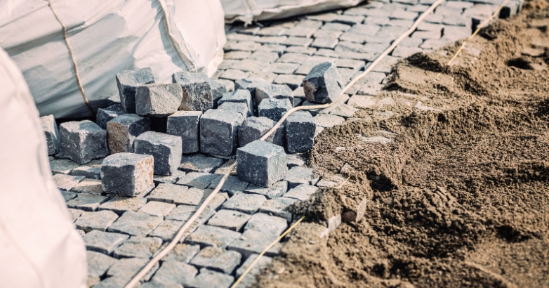 A pile of gravel and sand beside a bag, intended for a driveway cobblestone apron project.