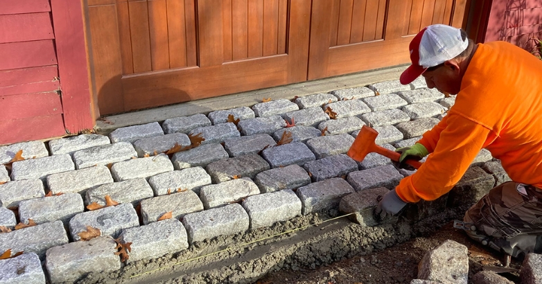 A man uses a hammer to install bricks for a cobblestone driveway apron, focusing on precise layout and alignment.
