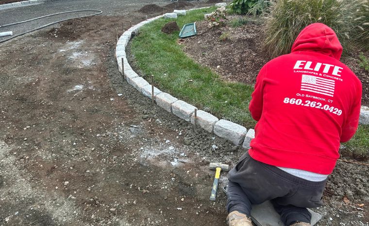 A man wearing a red hoodie is engaged in landscaping, focusing on custom cobblestone driveway edging in Clinton, CT.