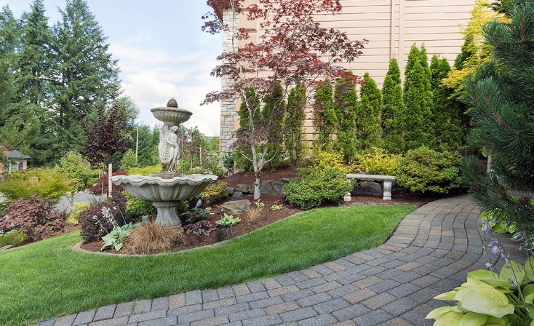 A brick pathway leads to a lush garden featuring a central fountain, showcasing the durability of Paver Walkway CT.