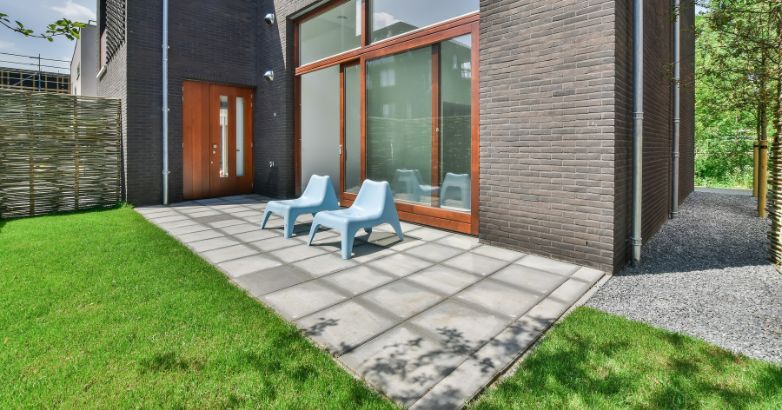 A modern backyard patio featuring large gray concrete tiles adjacent to a dark brick house with large glass doors.