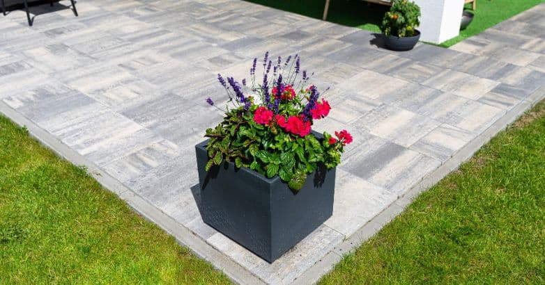Gray stone patio with a natural variegated pattern, bordered by green lawn and decorated with a colorful flower planter.