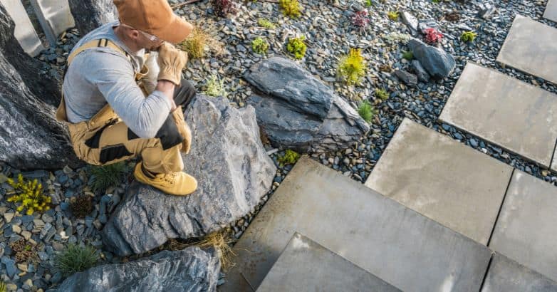 Landscaper inspecting an established stone walkway with large pavers, decorative gravel, natural boulders, and low-growing plants.