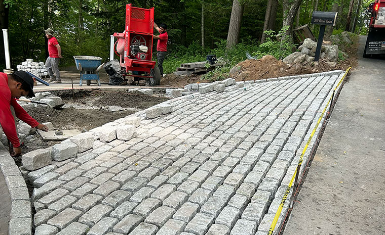 Workers installing cobblestone paver driveway in a wooded area.